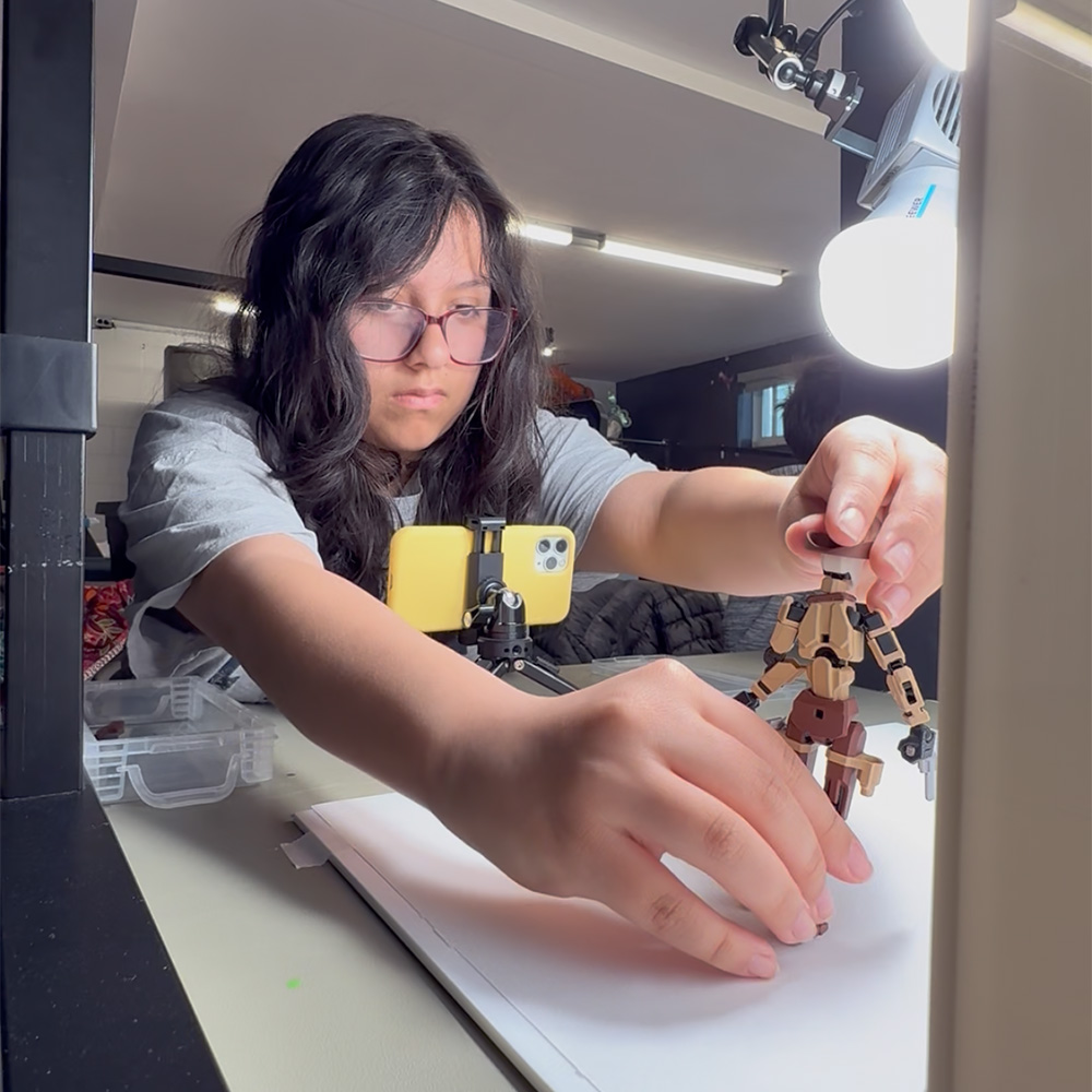 A student posing a wooden figure in front of a phone camera in a Storycraft Pioneers class for ages 11 to 14